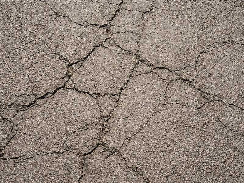 سطح آسفالت ترک‌خورده در نور روز با نمایش بافت، فرسودگی و جزئیات متریال